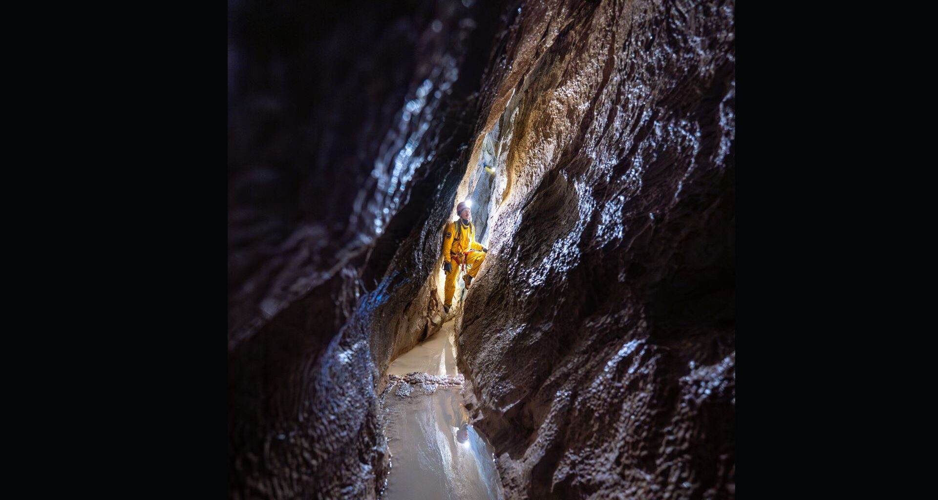 A man wearing a hard hat and a yellow jumpsuit looks up the wall of a narrow dark cave.