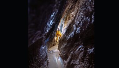 A man wearing a hard hat and a yellow jumpsuit looks up the wall of a narrow dark cave.