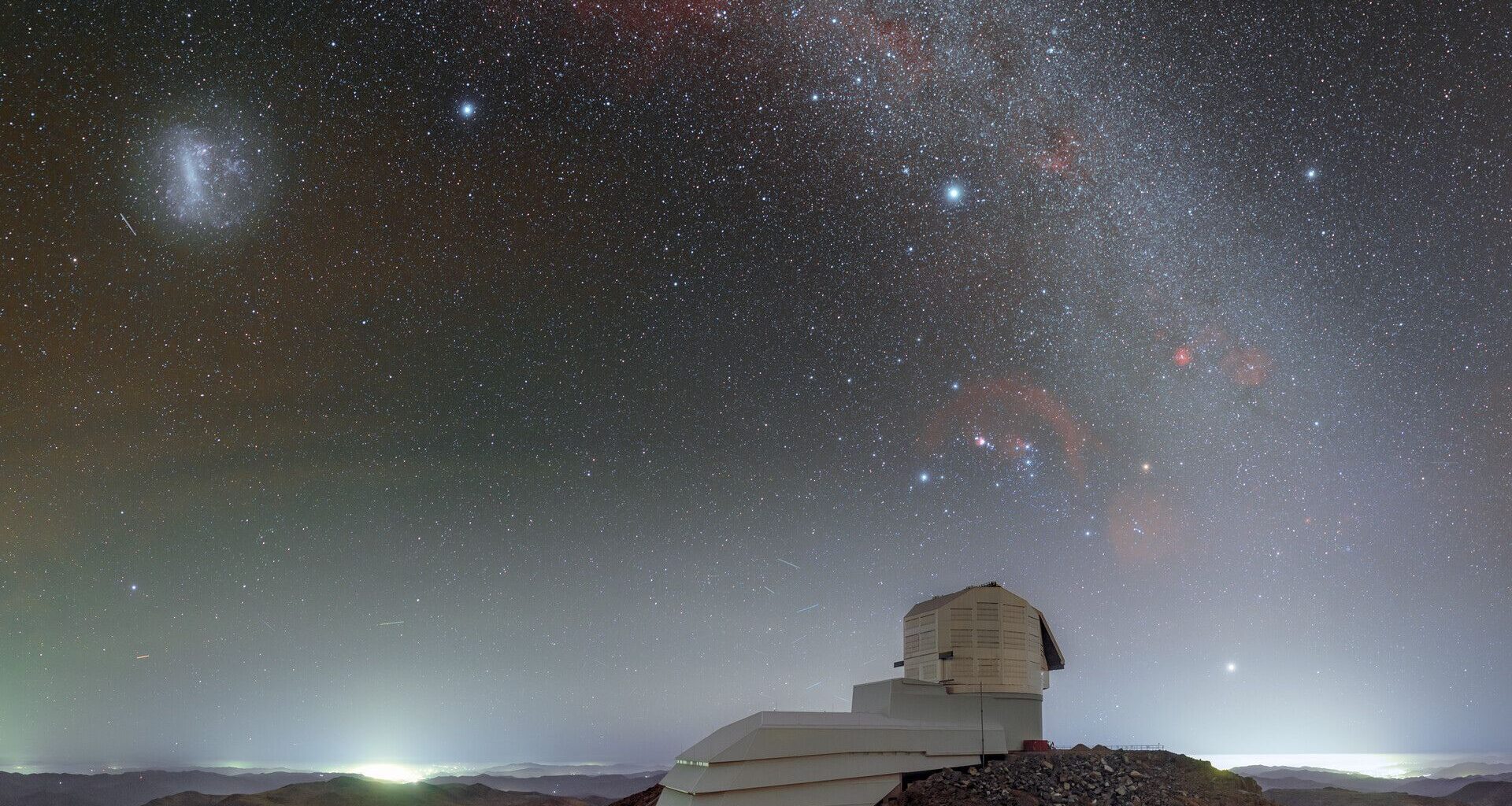 The domed roof of the Vera Rubin Observatory sits on a high ridge with a red and purple starry night sky above it with a glowing arch of the Milky Way seen in the heavens