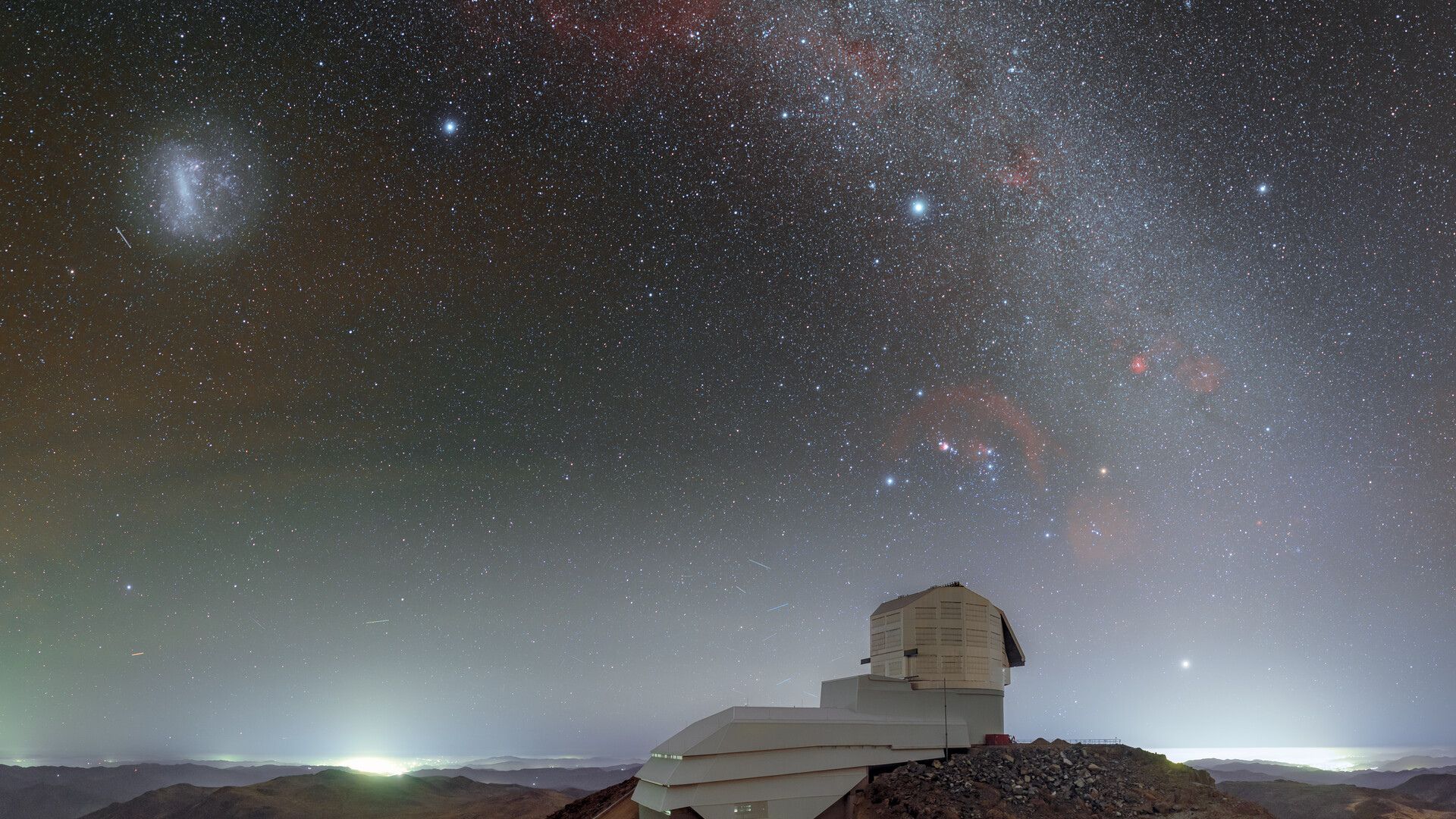 The domed roof of the Vera Rubin Observatory sits on a high ridge with a red and purple starry night sky above it with a glowing arch of the Milky Way seen in the heavens