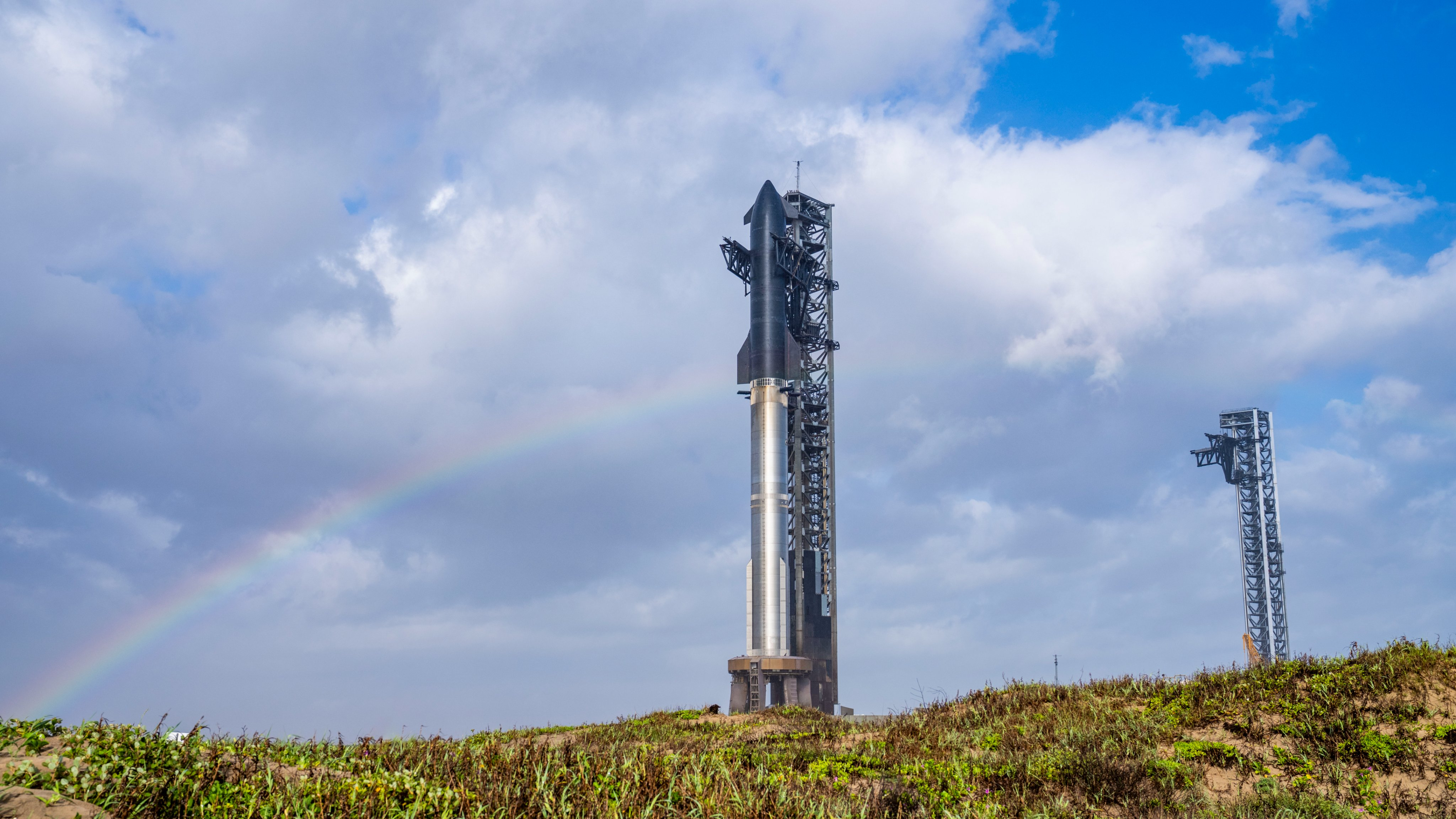 A giant rocket stands against bright cloudy blue sky crossed by a rainbow.