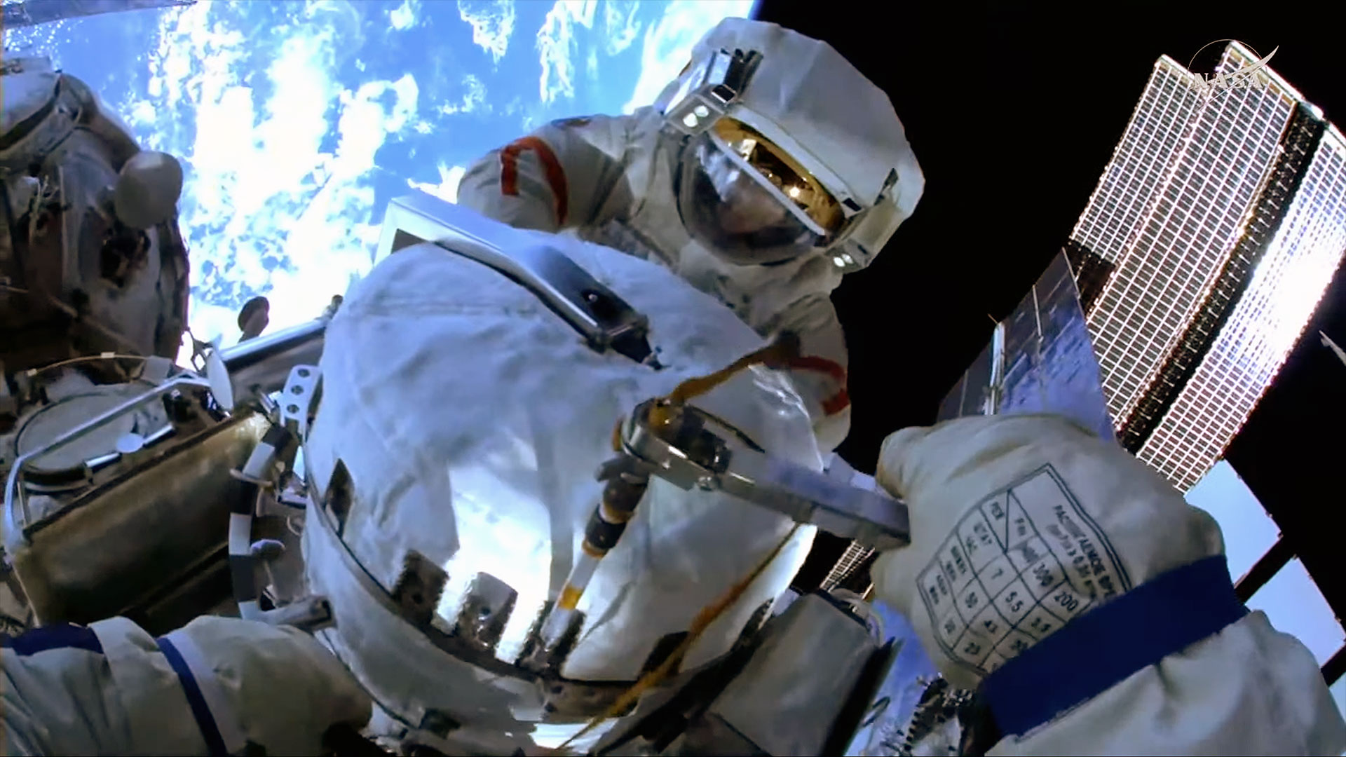 a camera mounted on a cosmonaut's spacesuit helmet captures a first-person view of another cosmonaut and Earth behind him while outside a space station.