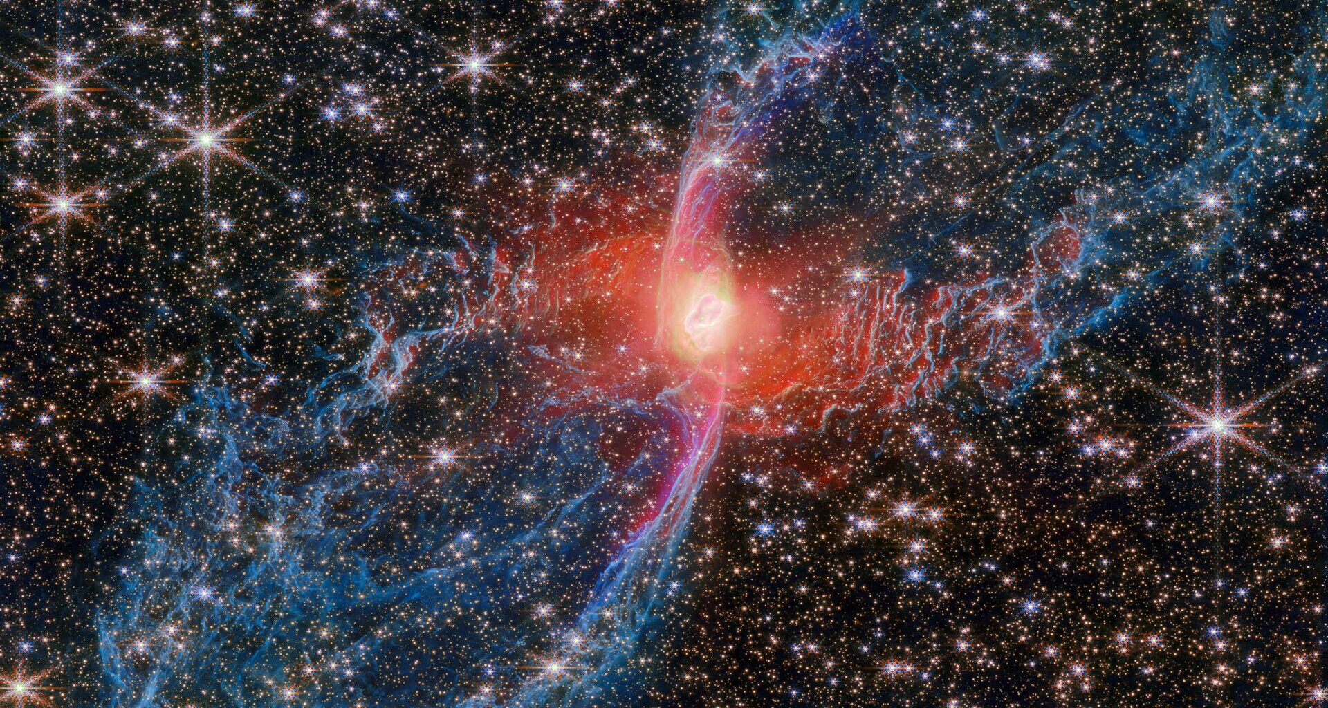 A large planetary nebula. The nebula&rsquo;s central star is hidden by a blotchy pinkish cloud of dust. A strong red light radiates from this area, illuminating the nearby dust. Two large loops extend diagonally away from the centre, formed of thin ridges of molecular gas, here coloured blue. They stretch out to the corners of the view. A huge number of bright, whitish stars cover the background, also easily visible through the thin dust layers.