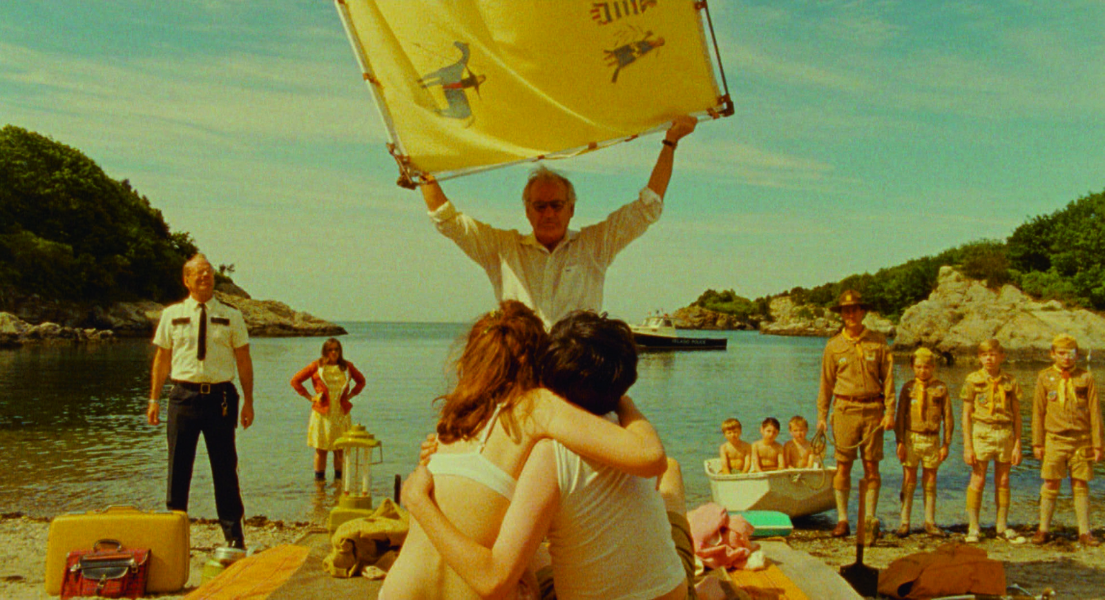 A beach scene with a couple embracing in the foreground, surrounded by several other people, a boat, and coastal scenery.