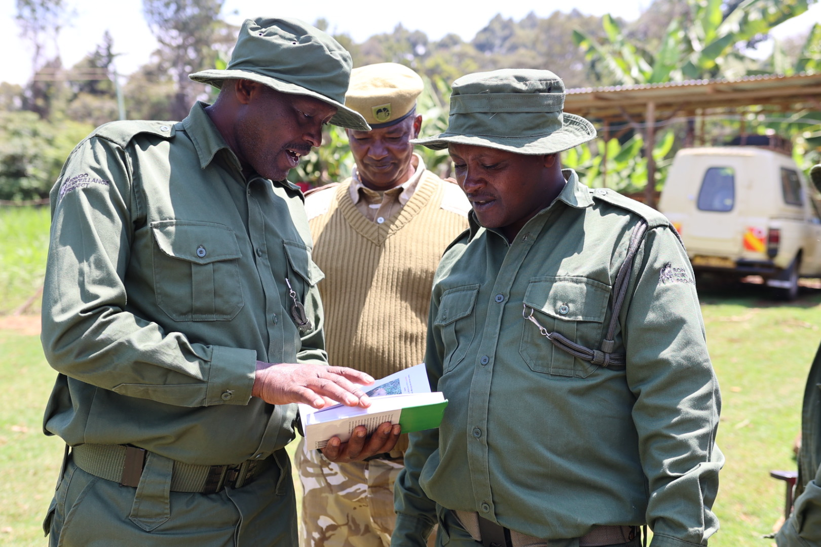 Three park rangers in uniform look at a book, with a vehicle in the background.