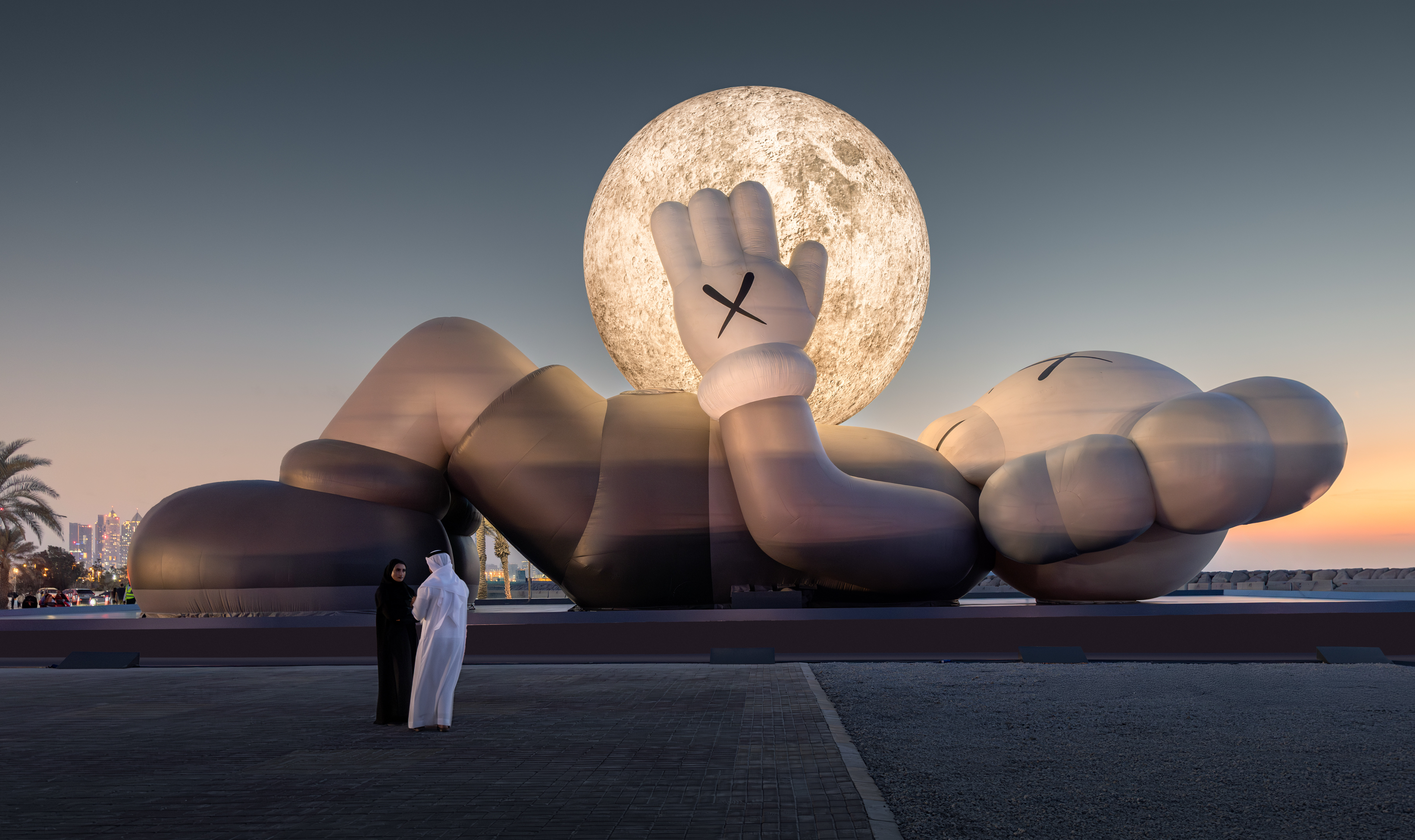 A 32-meter long KAWS Companion sculpture with an X on each hand rests on its side, lifting a large glowing moon, while two people observe it.