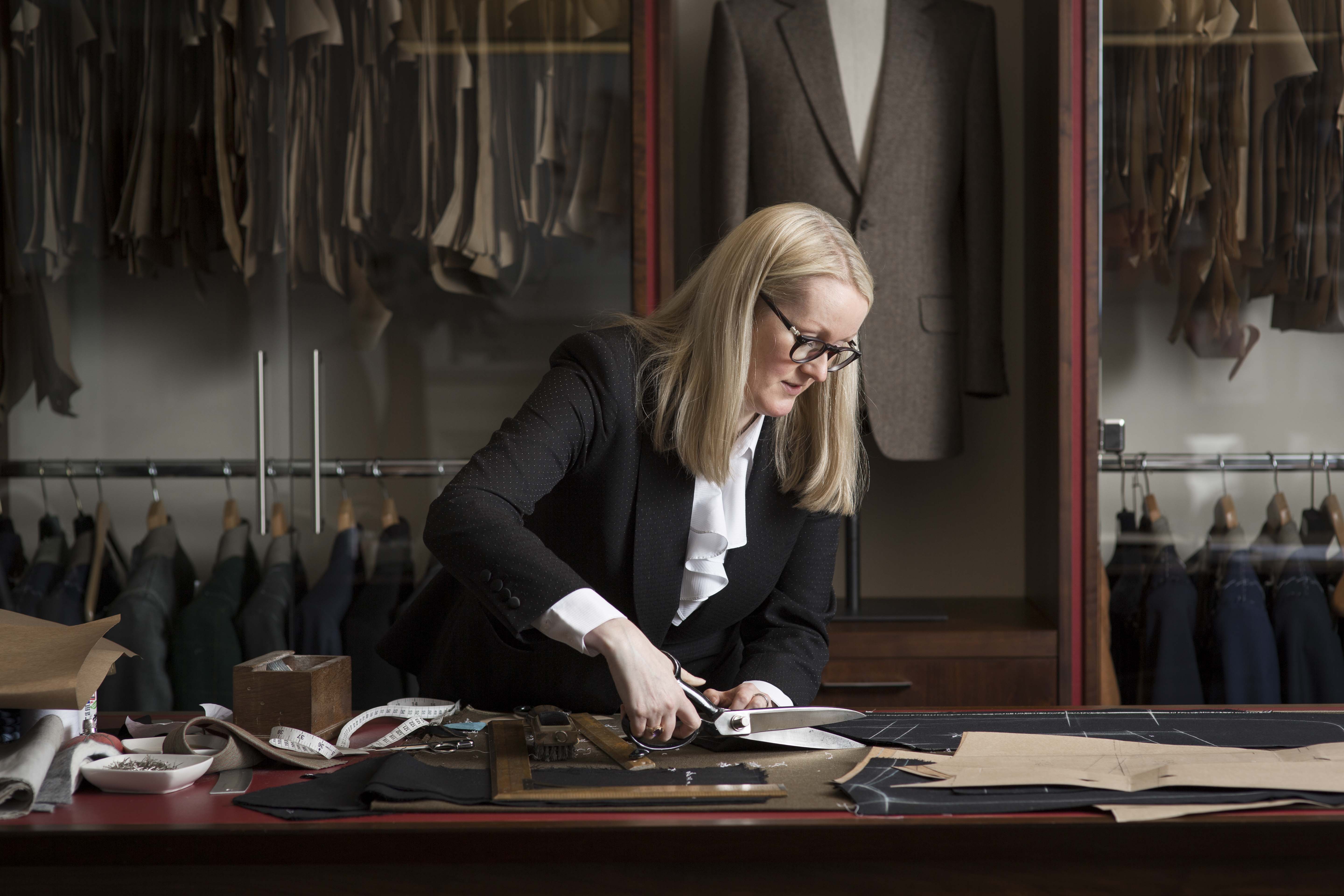 A woman tailor cutting fabric on a work table with various tools in a tailoring workshop.