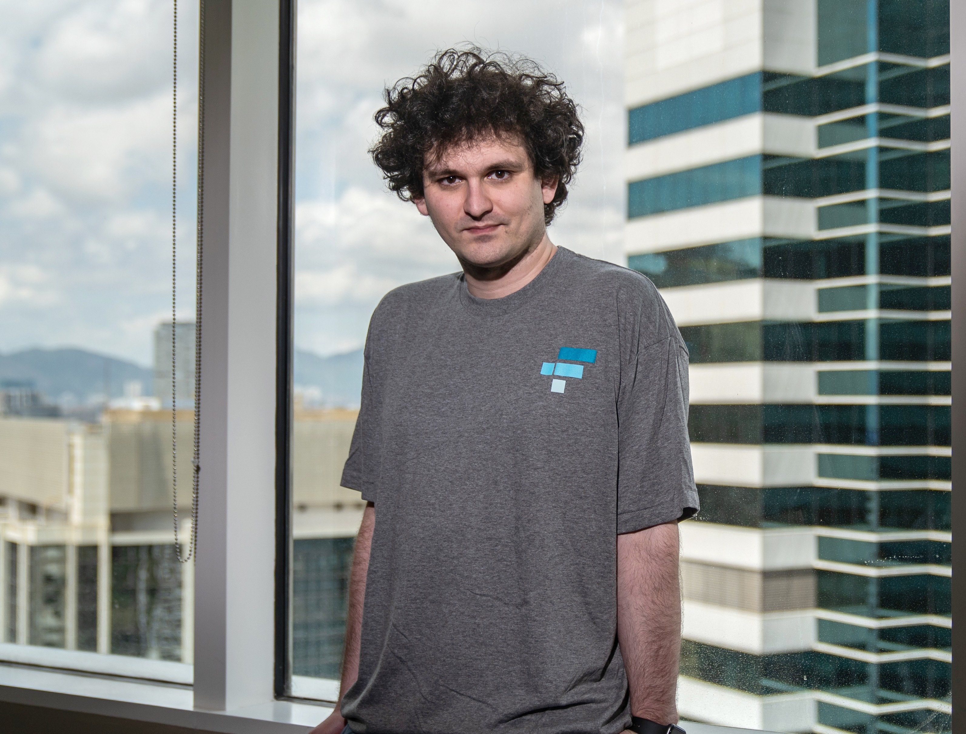 Sam Bankman-Fried, CEO of FTX, wearing a gray t-shirt with the FTX logo, looking at the camera, with a city skyline in the background.