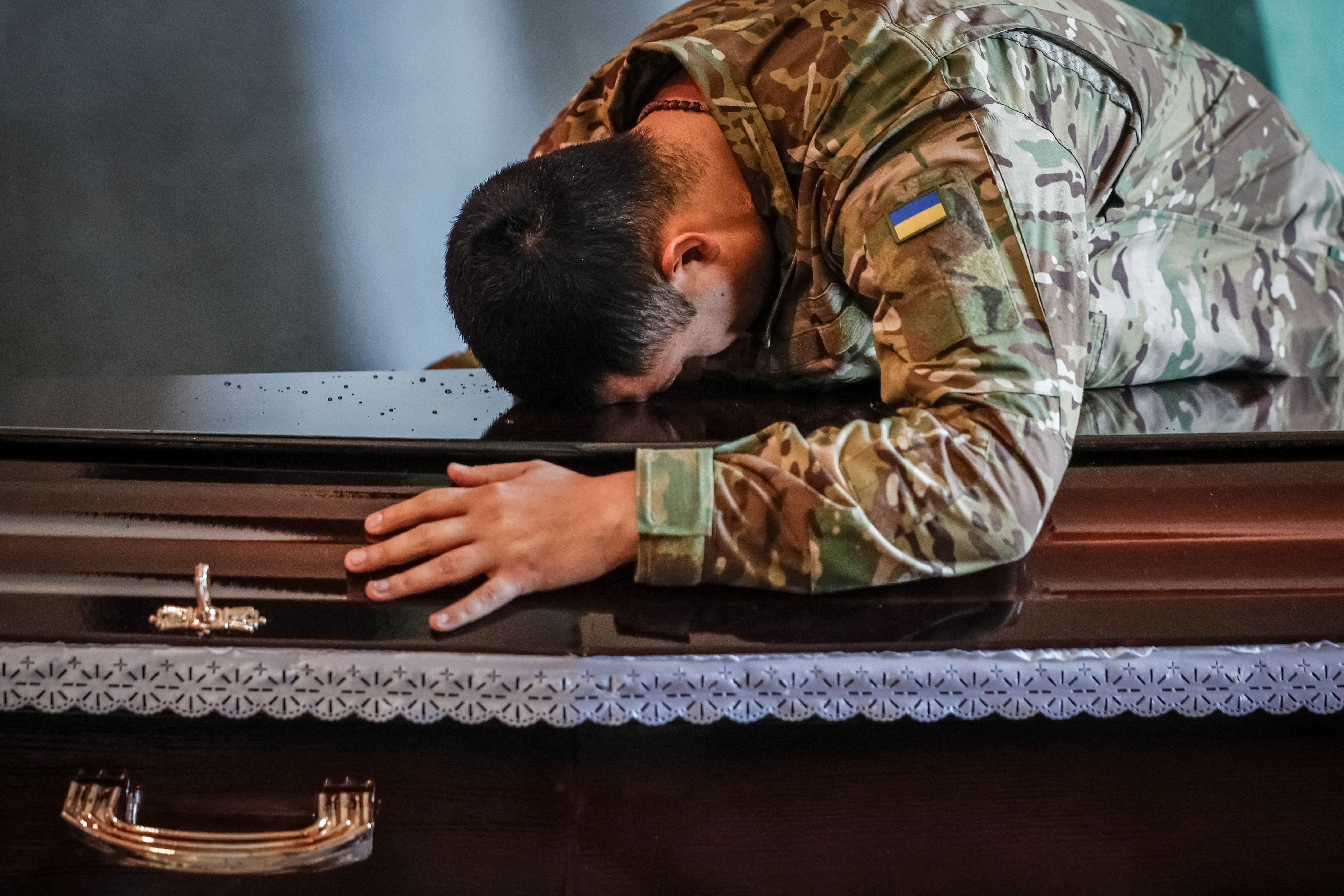 A soldier in camouflage uniform with a Ukrainian flag patch leans his head and hand on a dark casket during a farewell ceremony.