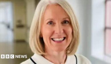 A woman wearing a light-coloured blouse with a black trim and a single dark button, standing indoors in a bright hallway with large windows.