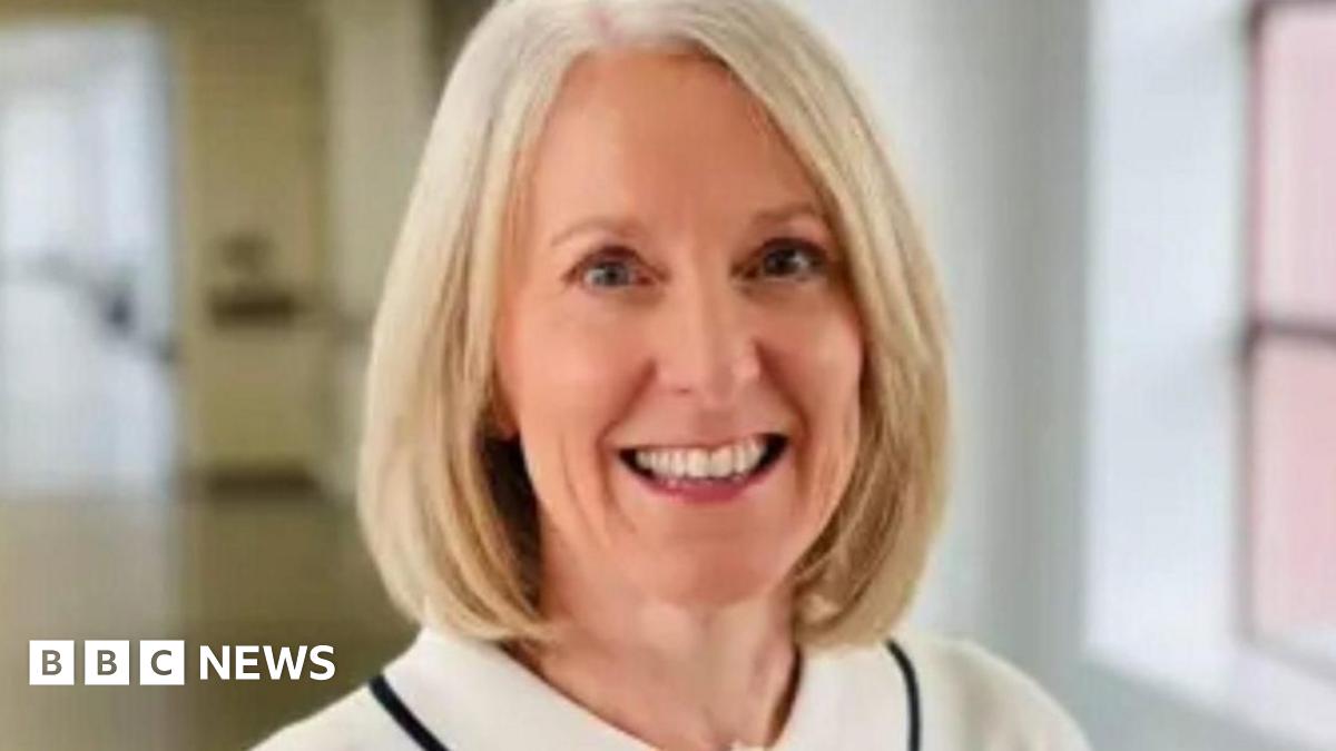 A woman wearing a light-coloured blouse with a black trim and a single dark button, standing indoors in a bright hallway with large windows.