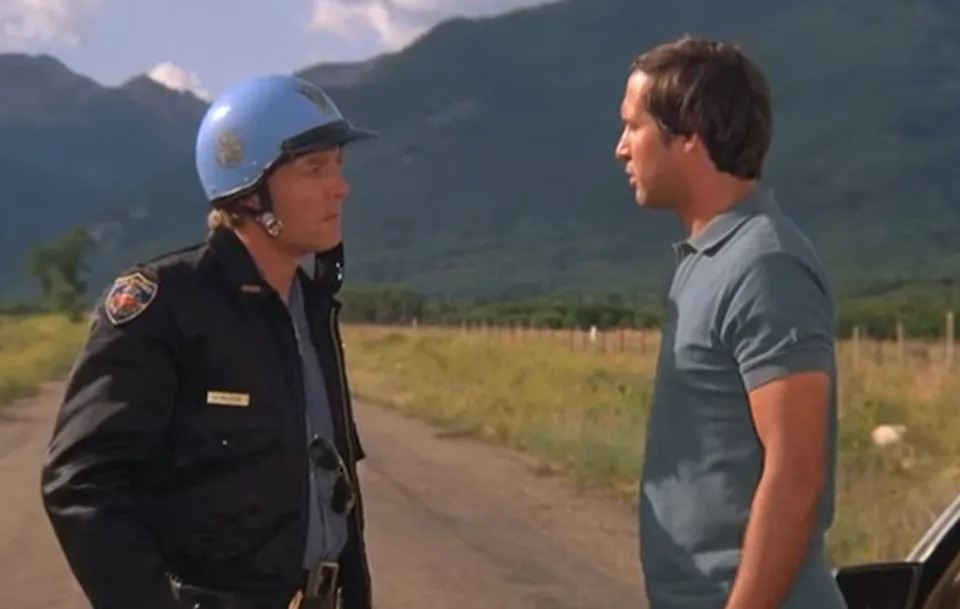 A person in a police uniform and helmet talks to a man in a casual polo shirt on a rural road with mountains in the background