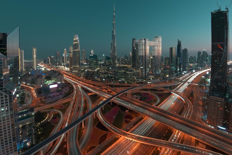 A vibrant cityscape at dusk shows a network of illuminated highways and skyscrapers, including the tall Burj Khalifa at the center, under a clear teal sky in Dubai.