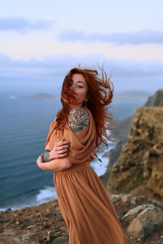 A woman with long red hair wearing a flowing tan dress stands on a rocky cliffside by the ocean, her hair blowing in the wind. She has a large floral tattoo on her shoulder and gazes confidently at the camera.