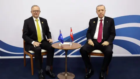 Getty Images Two men in suits sit in chairs. Between them is a round table with two small flags: one Australian and the other Turkish