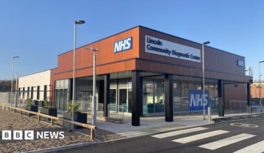A modern building with open windows on the bottom with a brown upper floor exterior and the words NHS and Lincoln Community Diagnostic Centre written on it.