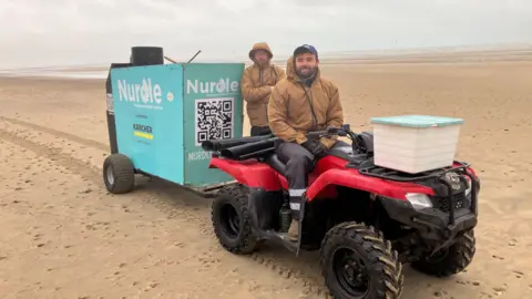 Sam Dixon-French/BBC Josh Beech sits on a quad bike on Camber Sands, with Dominic Manning standing behind him.