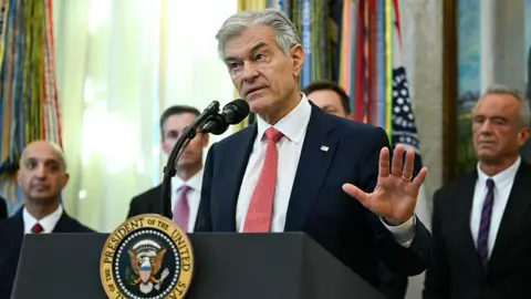 Getty Images Medicare and Medicaid Administrator Mehmet Oz speaks in the Oval Office during an event about weight-loss drugs at the White House in Washington, DC on November 6, 2025. He is speaking at a podium that features the seal of the President of the United States. His hair is short and grey, and he is wearing a black suit with a red tie.