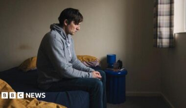 A wearing a grey hood and jeans is sitting on a bed.  He has a sad look on his face and he is looking down at the floor, with his hands clenched.
