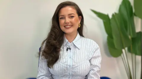 A woman with long brown hair, wearing a pale blue striped shirt is sitting on a blue chair with a large green plant beside her. She is smiling into camera