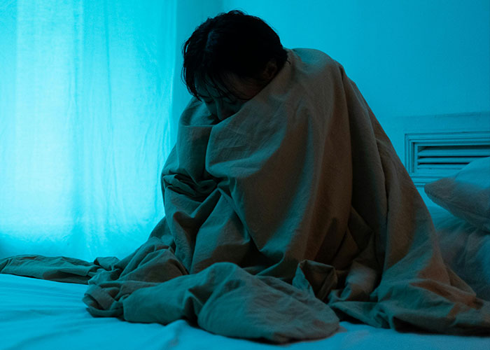 Person wrapped in a blanket sitting on a bed in a dimly lit room, reflecting the emotional challenges of psychiatric institution work.