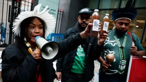 AFP via Getty Images Wakrewa Krenak, left, who lives in Conselheiro Pena, 400km from Mariana, holds a bottle of the contaminated water retrieved from the river Doce, as she speaks in a loud speaker during a victim gathering outside the High Court in London, on 21 October 2024.
