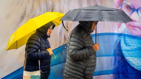 PA Media A man and a woman walk through a city centre past a billboard poster. The woman has a yellow umbrella and the man a black one. Both are wearing winter coats with the hoods up.