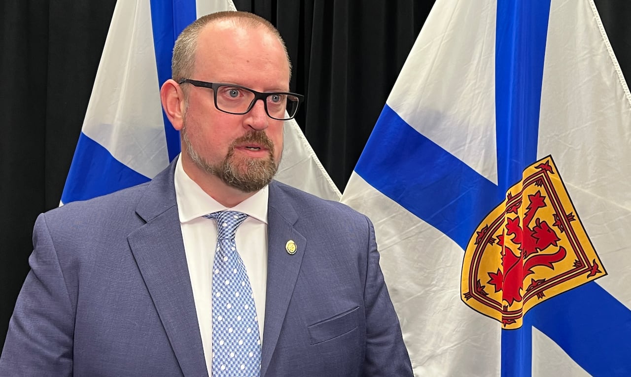 A man in a suit and tie with glasses stands by a Nova Scotia flag.
