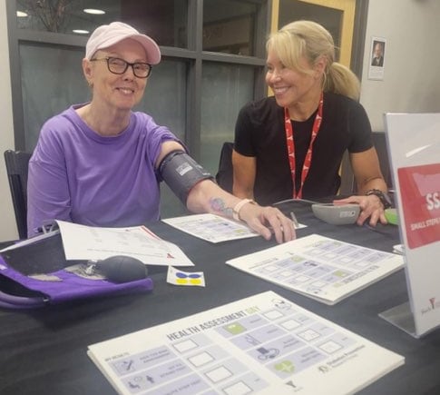 Two women sitting at a table smiling. One is wearing an arm cuff for blood pressure and heart rate testing.