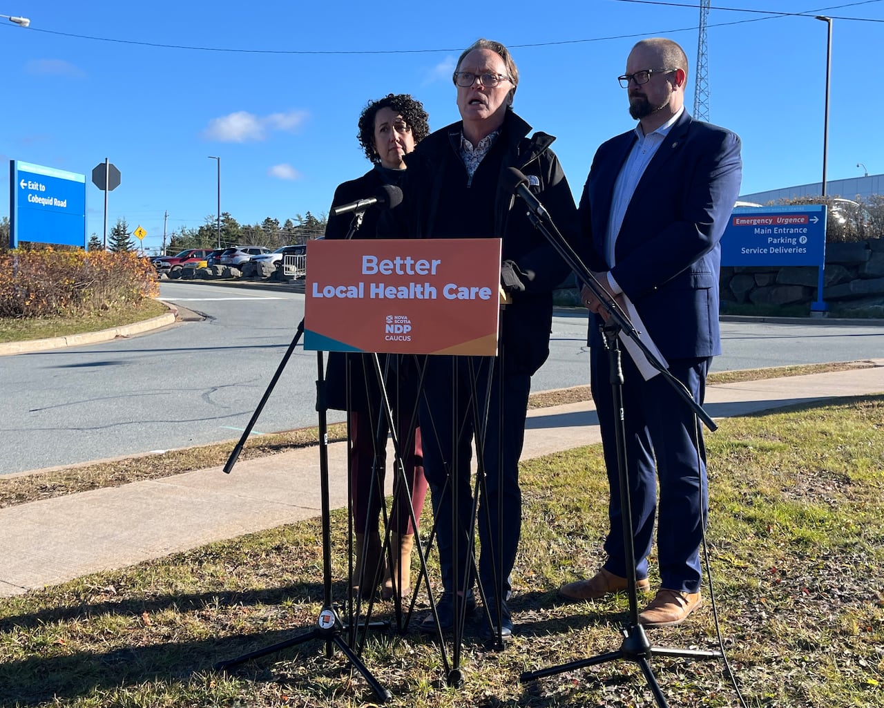 Three people stand behind a podium in front of a hospital.