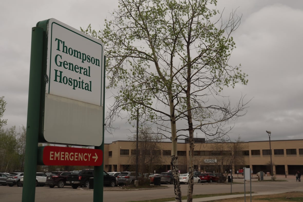 A tree stands beside a white sign that says 'Thompson general hospital' and has a brown brick building with a parking lot full of cars.