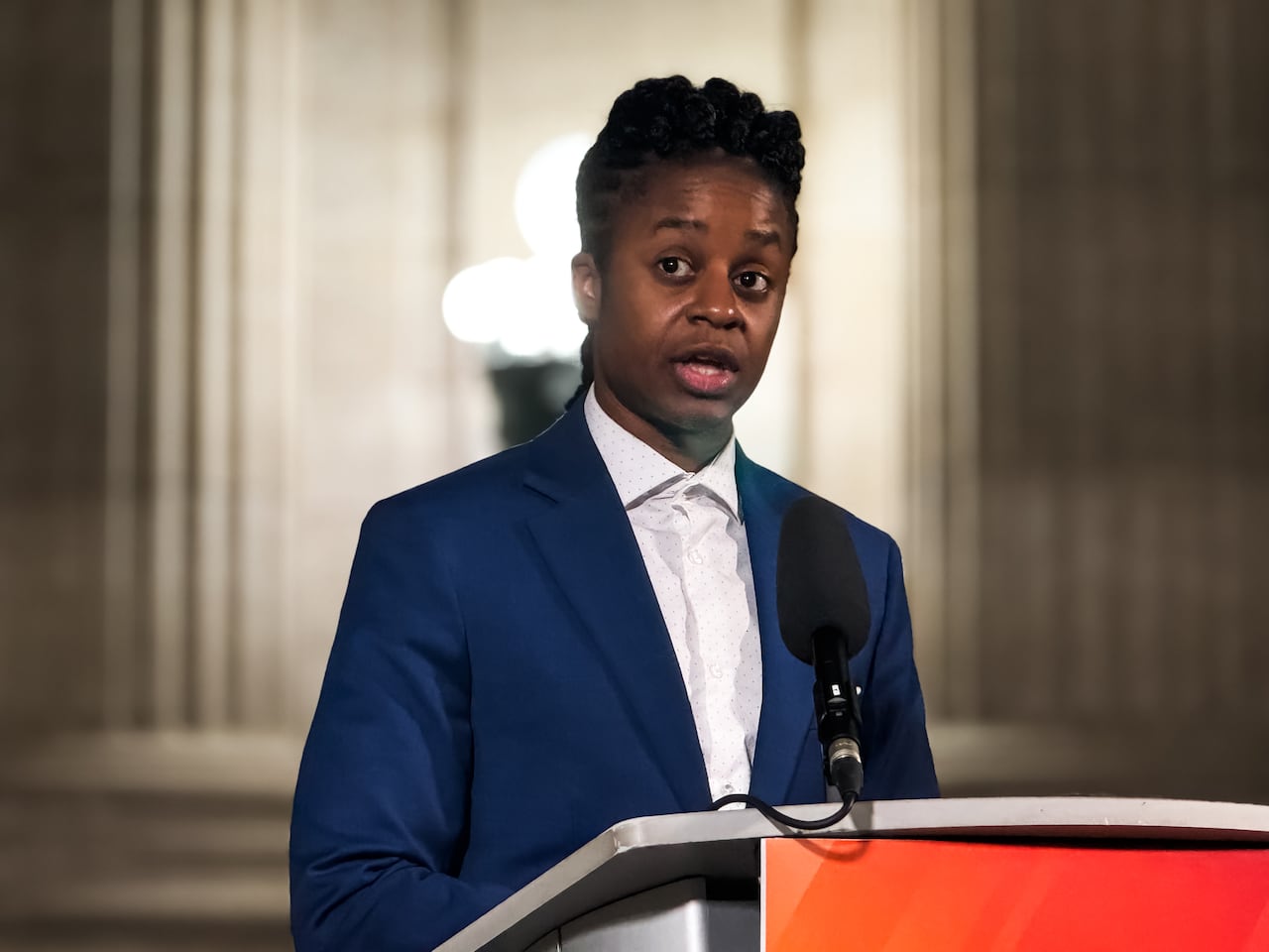 A person in a blue blazer and white t-shirt opens their mouth as they talk.