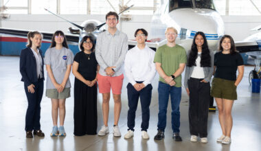 A group of eight people stand together inside a hangar with a somewhat shiny floor. In the background is a small white plane with a blue stripe, and large windows behind that.