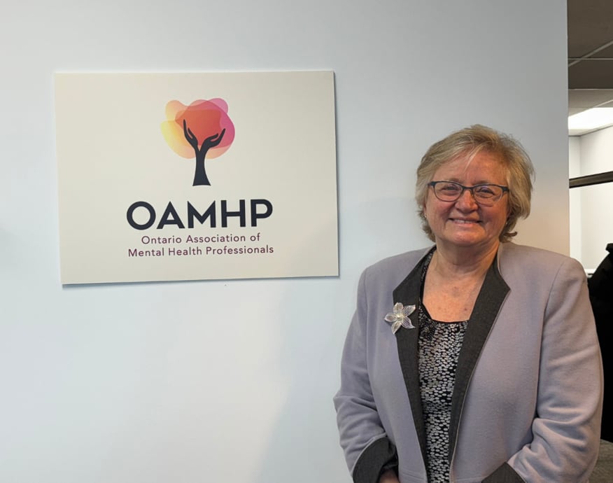 A woman wears a grey blazer on a black and white spotted blouse with OAMHP on a sign beside her. 
