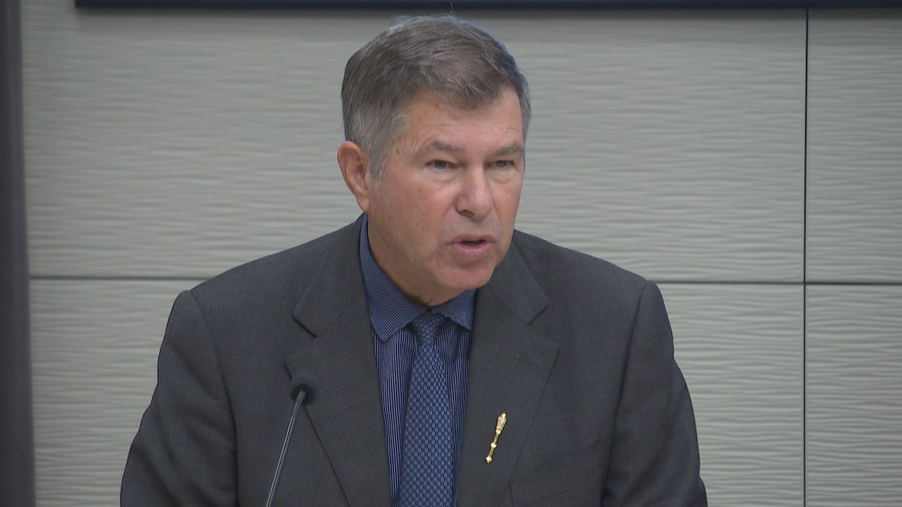 A man with short, greying hair, wearing a blazer, dress shirt and tie, speaking into a microphone.