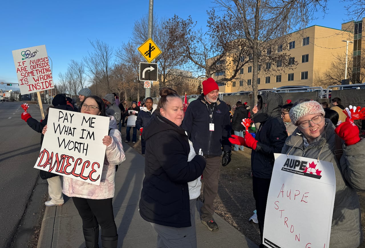 Health-care workers dressed in coat and hats hold signs along the side of the road; one reads "Pay me what I'm worth, Danielle."