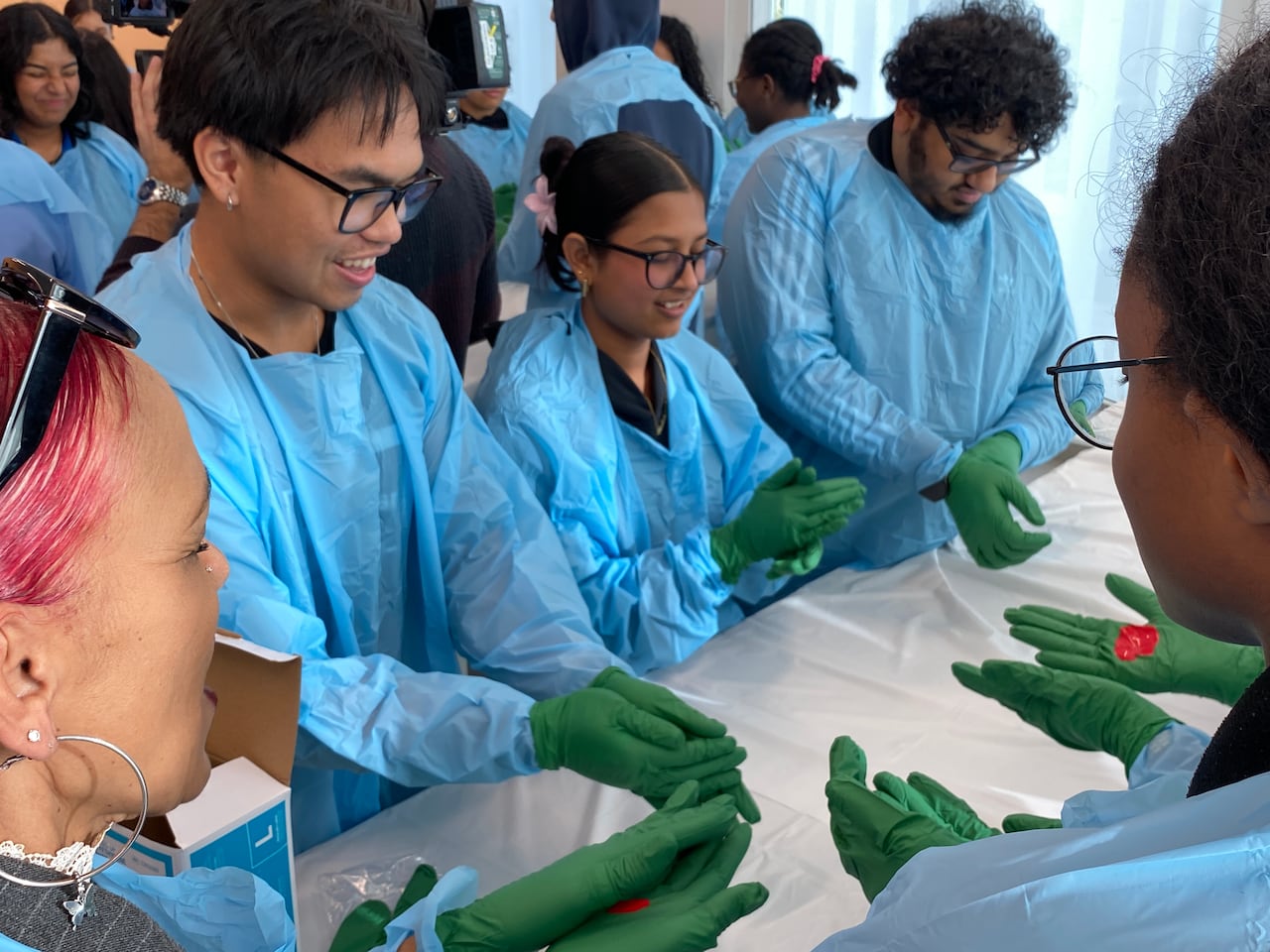 Students and adults donning disposable green gloves and blue plastic gowns close their eyes as they rub their hands together with red paint. 