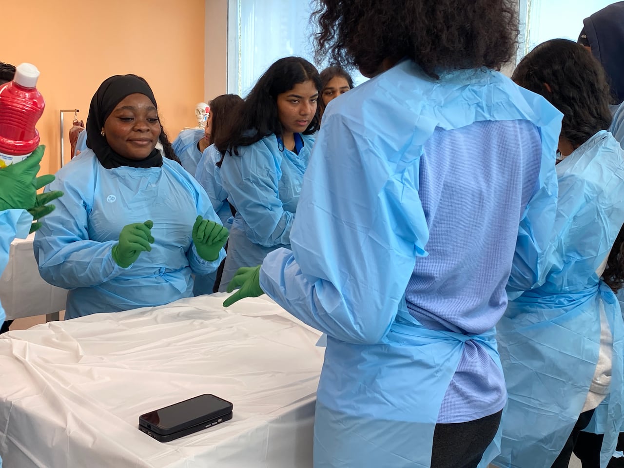Students in green plastic gloves and blue plastic protective gowns over their clothes stand around a table covered with a white tablecloth.