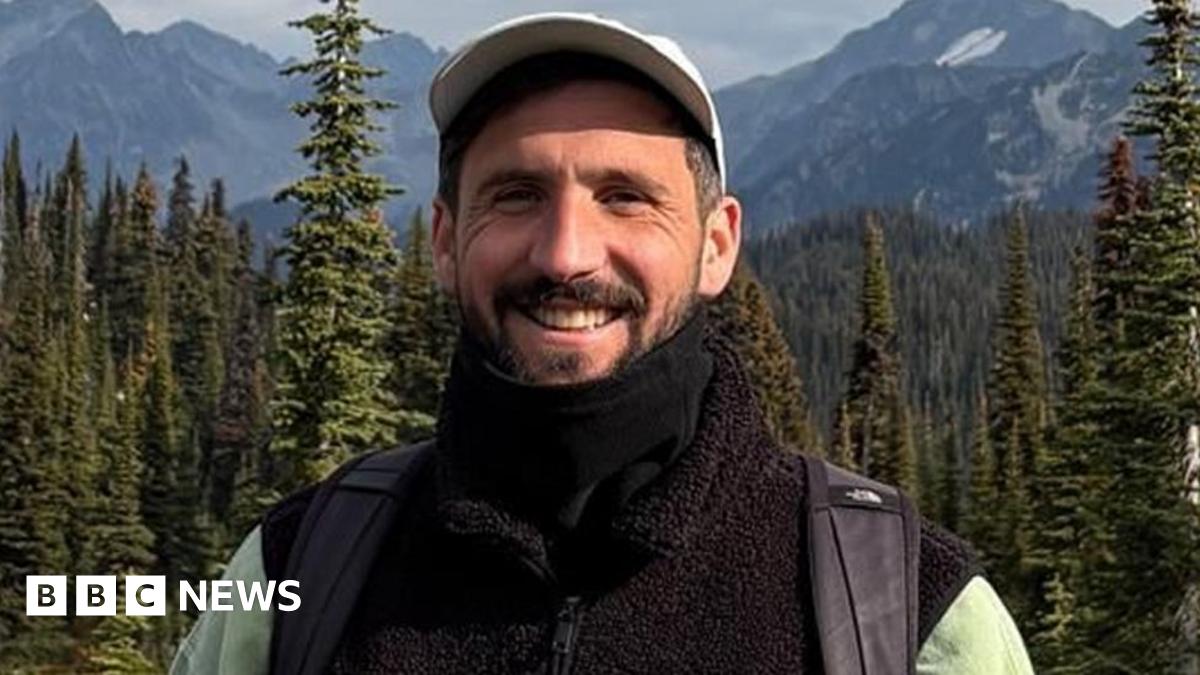 Oliver Gooch standing smiling in front of trees and mountains smiling. He has dark hair and a beard and is wearing a white baseball cap and black fleece and scarf