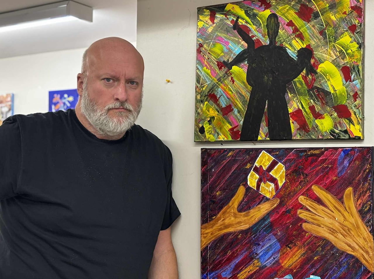 A man stands next to two colourful paintings mounted to the wall. 