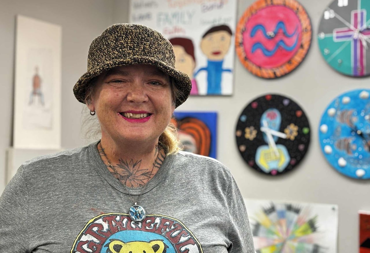 A woman smiles at the camera. Behind her are four colourful hand-painted clocks mounted on the wall