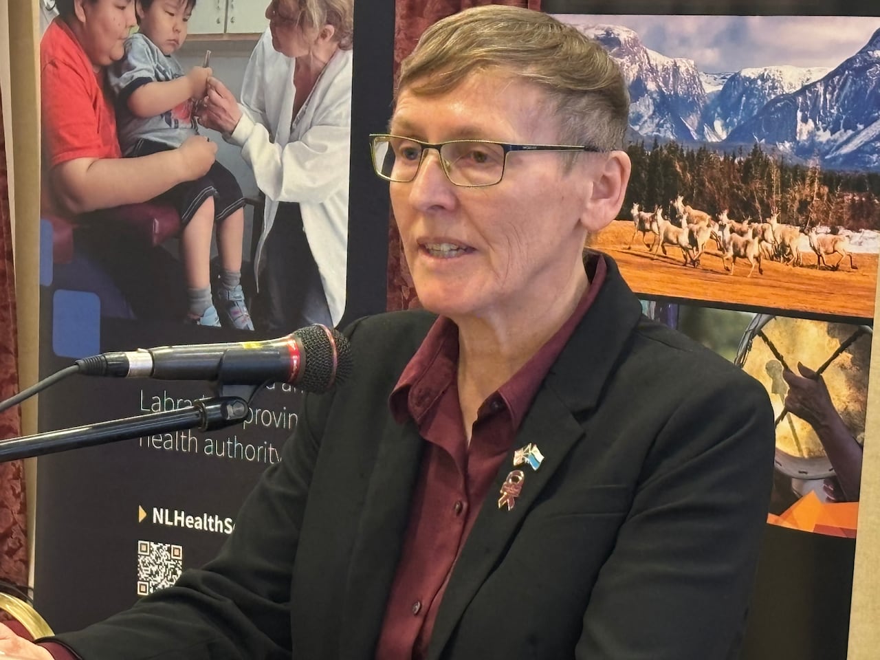 A woman with short brown hair and glasses sits at a microphone.