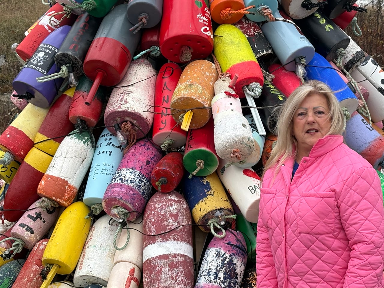 A white lady with blonde hair, wearing a pink jacket, standing in front of a Christmas tree decorated using colourful fishing buoys.