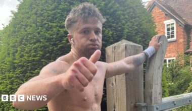 Tommy, a young man, has his top off and is doing a thumbs up at the camera with a tired look on his face. He is resting his other hand on a wooden gate by a house. He has several tattoos, including a cross on his sternum.