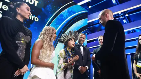 PA Media William on stage with five others, including a woman in an all-black dress, a young woman with long curly blonde hair wearing a sleeveless white dress, a young person wearing a feathered headdress and white patterned top, and two men in tuxedos.