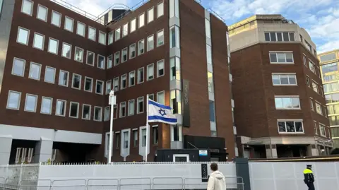 PA Israel embassy in Dublin, a large brown building with lots of windows, a white flag with blue star and banners is blowing in front of building.