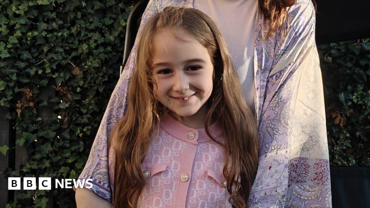 A girl with long brown hair is standing in a garden. The child is wearing a pink cardigan with a geometric pattern and blue jeans. A woman is standing behind the girl. She is wearing a long patterned kimono-style garment over a beige top. Behind them is a fence covered in green ivy.