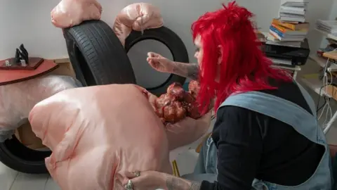 London Museum A woman with bright red hair, a black top and denim dungarees works on a peach coloured piece of art which involves a silk-like peach-coloured material and pearls. 
