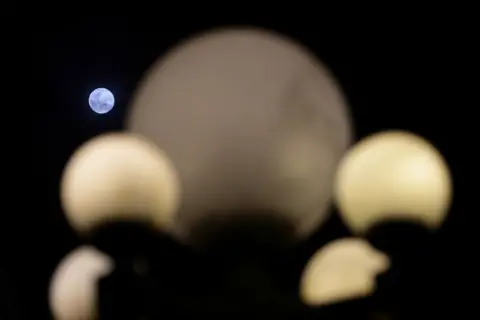 Suhaimi Abdullah / NurPhoto via Getty Images Circles of the streetlamp shine in the foreground while the Moon rises against a dark night sky in Singapore on Wednesday.