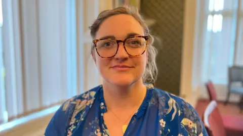 A person wearing a blue floral-patterned blouse sits indoors in a room with tall vertical blinds, patterned wallpaper, and upholstered chairs. The background includes a radiator and framed artwork